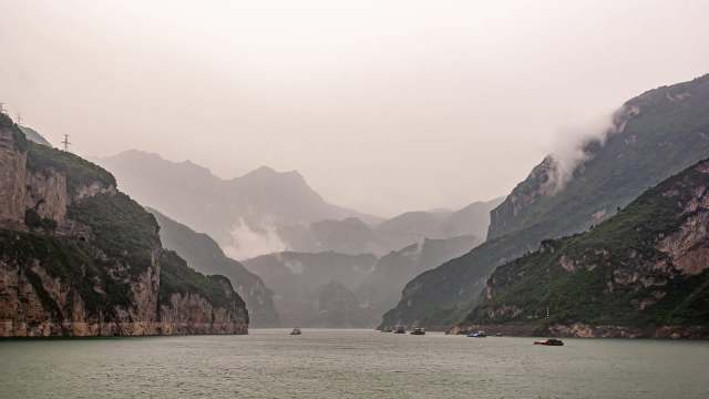 A misty landscape of mountains with a river, featuring boats amidst lush green cliffs.