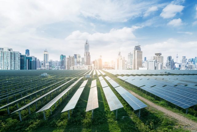 A solar panel farm in the foreground with a modern city skyline in the background under a bright sky.