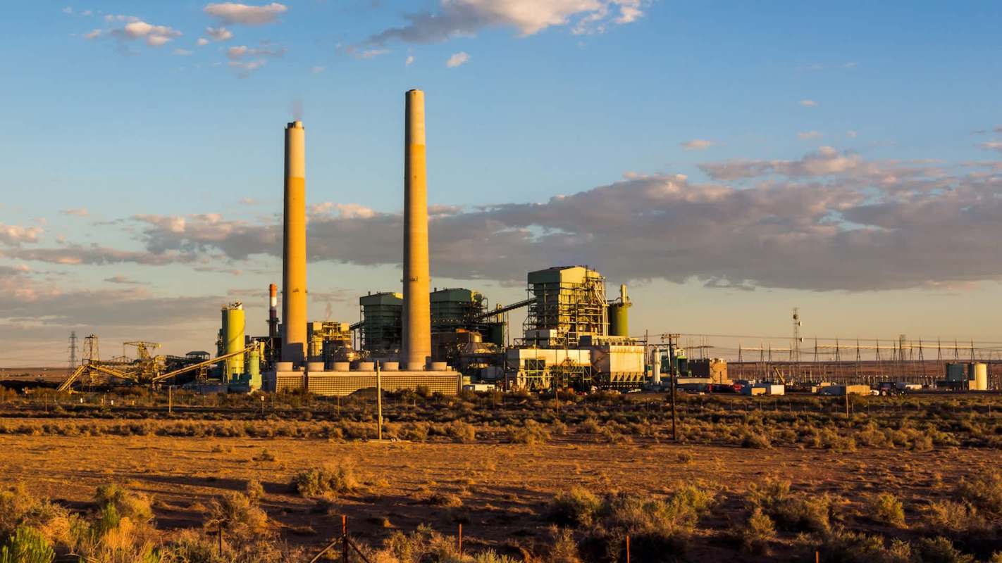 A power plant with two tall smokestacks and industrial structures under a blue sky with scattered clouds.