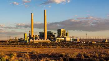 A power plant with two tall smokestacks and industrial structures under a blue sky with scattered clouds.