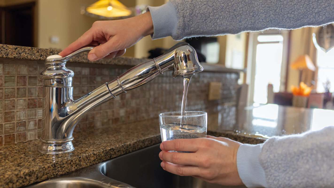 A hand turns on a faucet, pouring water into a glass on a granite countertop in a bright kitchen.