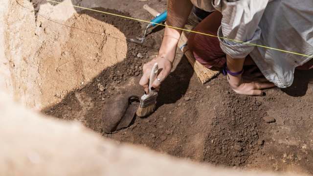 A person carefully brushes dirt away from a pottery fragment during an archaeological dig.