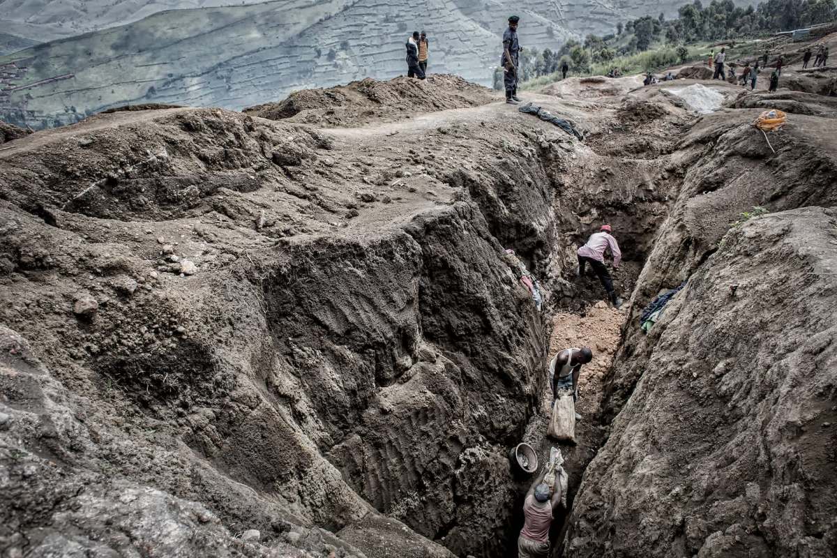 Workers dig deep trenches in a rugged landscape, with mountains and hills visible in the background.