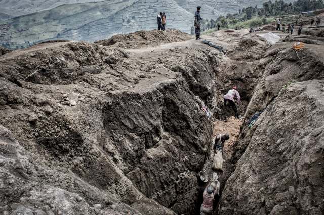 Workers dig deep trenches in a rugged landscape, with mountains and hills visible in the background.