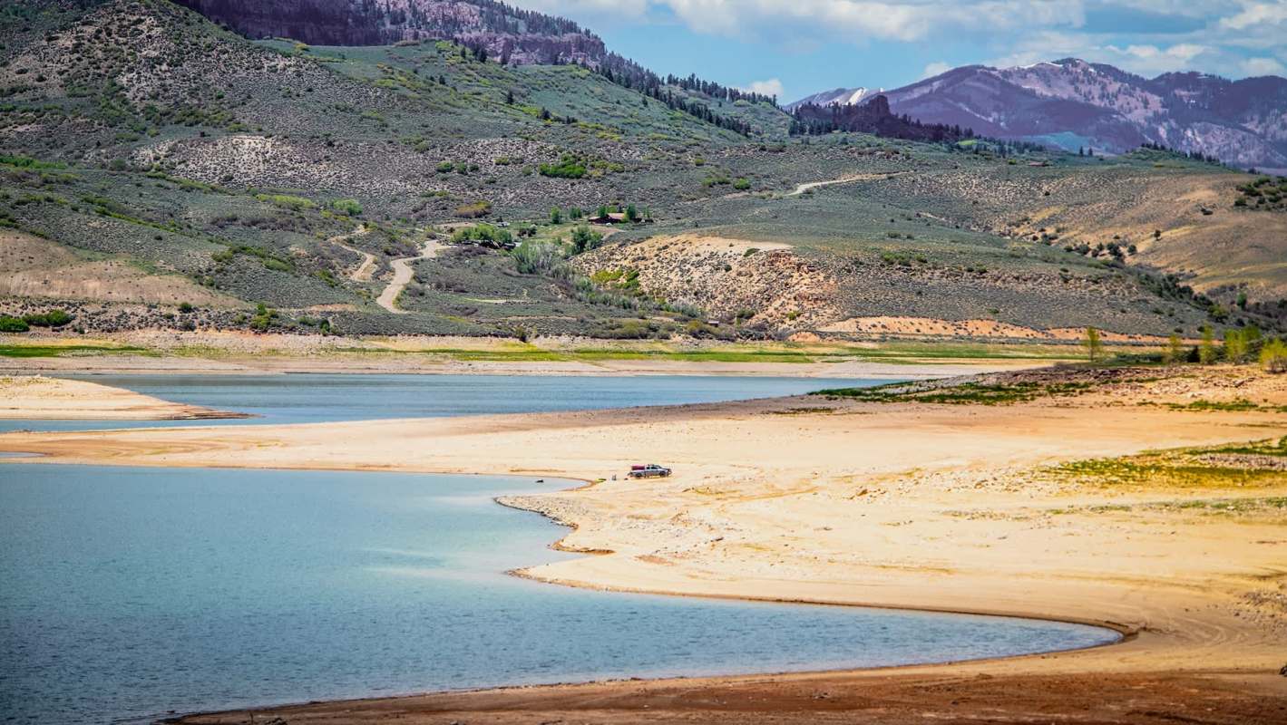 A serene lake bordered by sandy shores and rolling green hills under a partly cloudy sky.
