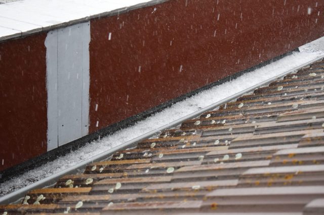 Snow or hail falls on rooftops, creating a white accumulation along the edges and a gray sky above.
