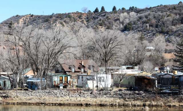 A cluster of rustic homes and bare trees is nestled near a calm body of water against a mountainous backdrop.