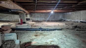 A dimly lit basement with exposed beams, pipes, and a concrete floor featuring construction materials scattered around.