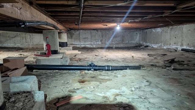 A dimly lit basement with exposed beams, pipes, and a concrete floor featuring construction materials scattered around.