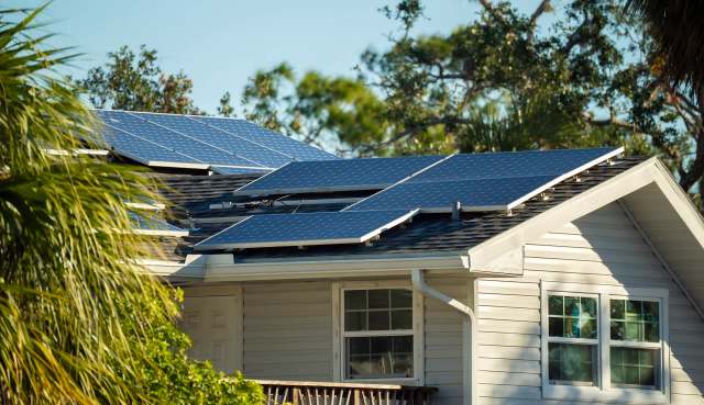 A home with solar panels installed on its sloping roof, surrounded by trees and greenery.