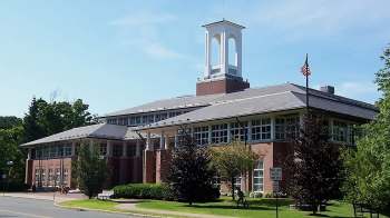 A three-story brick building with a clock tower and American flag surrounded by greenery on a clear day.