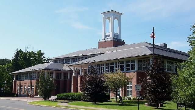 A three-story brick building with a clock tower and American flag surrounded by greenery on a clear day.