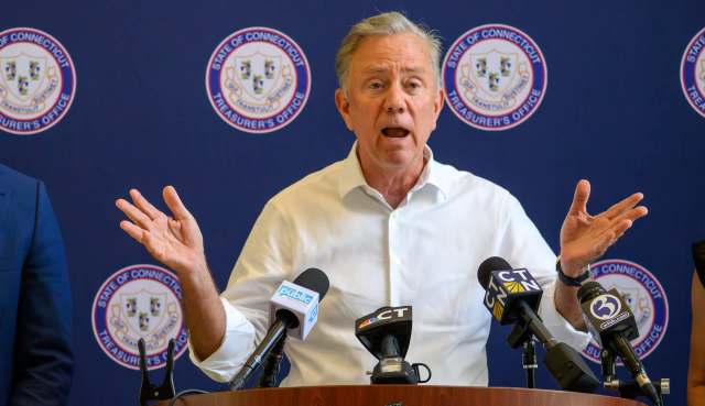 Connecticut Gov. Ned Lamont gestures while speaking at a podium surrounded by microphones and a blue backdrop.