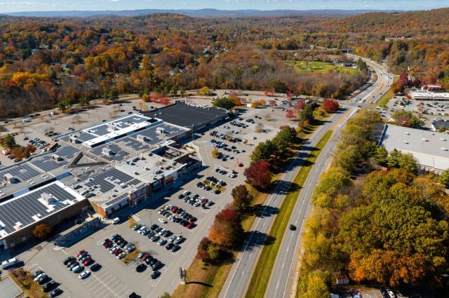 Aerial view of a shopping center surrounded by vibrant autumn foliage and a nearby highway.