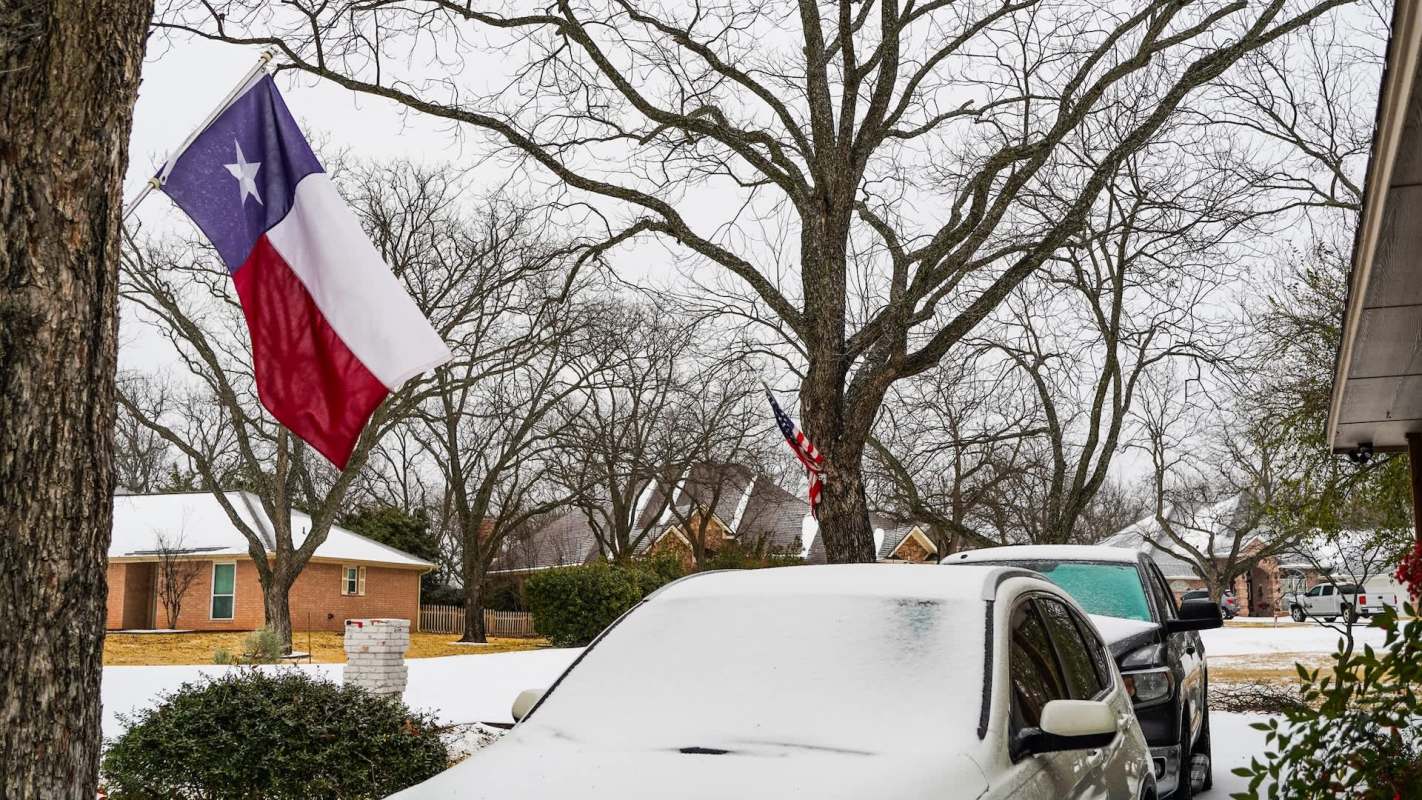 A Texas flag and an American flag hang near snow-covered cars in a suburban neighborhood.