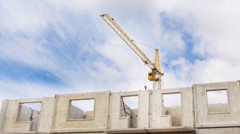 A yellow construction crane operating above partially constructed concrete walls under a blue sky with clouds.