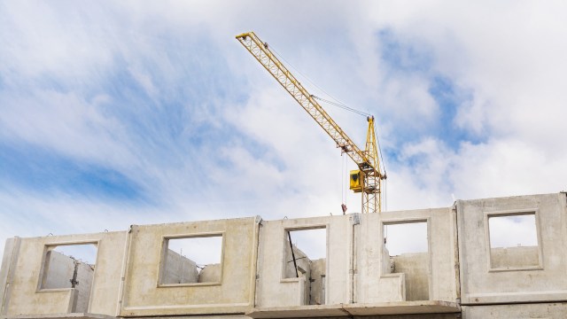 A yellow construction crane operating above partially constructed concrete walls under a blue sky with clouds.