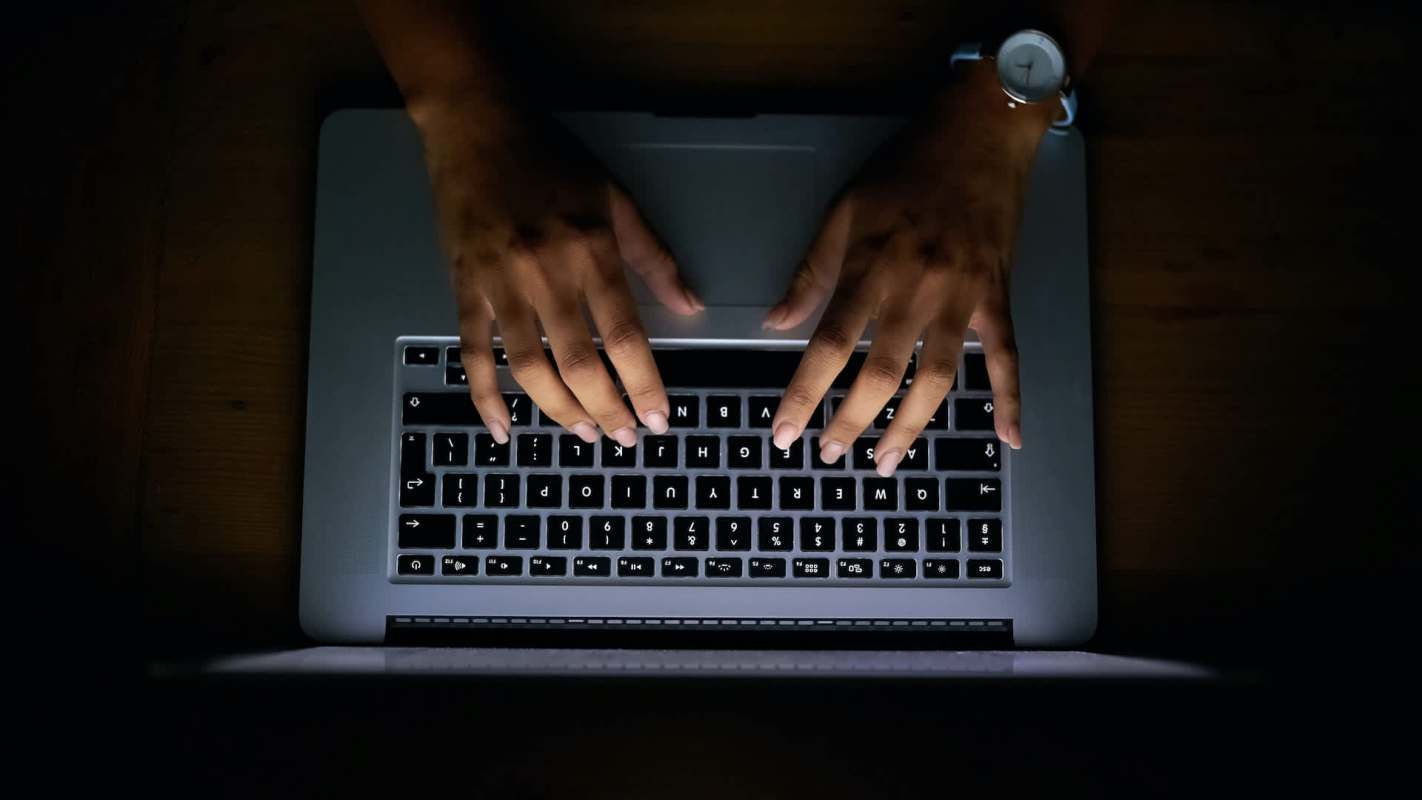 A person's hands typing on a laptop keyboard in a dimly lit environment.