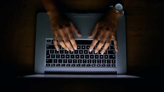 A person's hands typing on a laptop keyboard in a dimly lit environment.