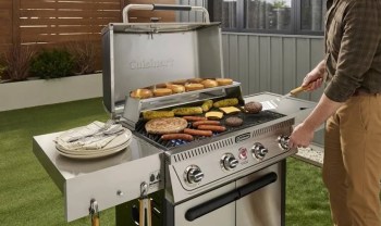 A person grilling various foods, including burgers, hot dogs, and corn, on a stainless steel barbecue in a backyard.