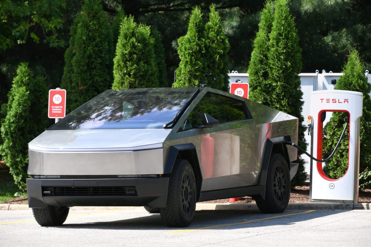 A futuristic Tesla Cybertruck charging at a supercharger station surrounded by green shrubs.