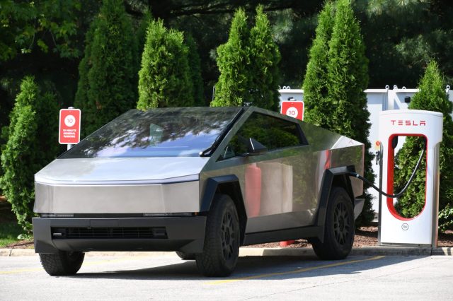 A futuristic Tesla Cybertruck charging at a supercharger station surrounded by green shrubs.