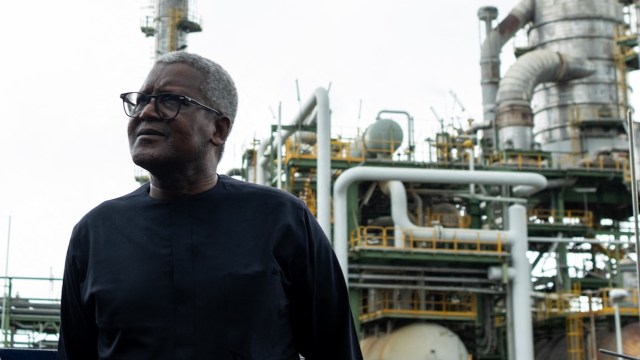 Aliko Dangote stands in front of an industrial plant with numerous pipes and structures.