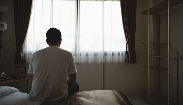 A person sitting on a bed, viewed from behind, gazes out a window with sheer curtains in a softly lit room.