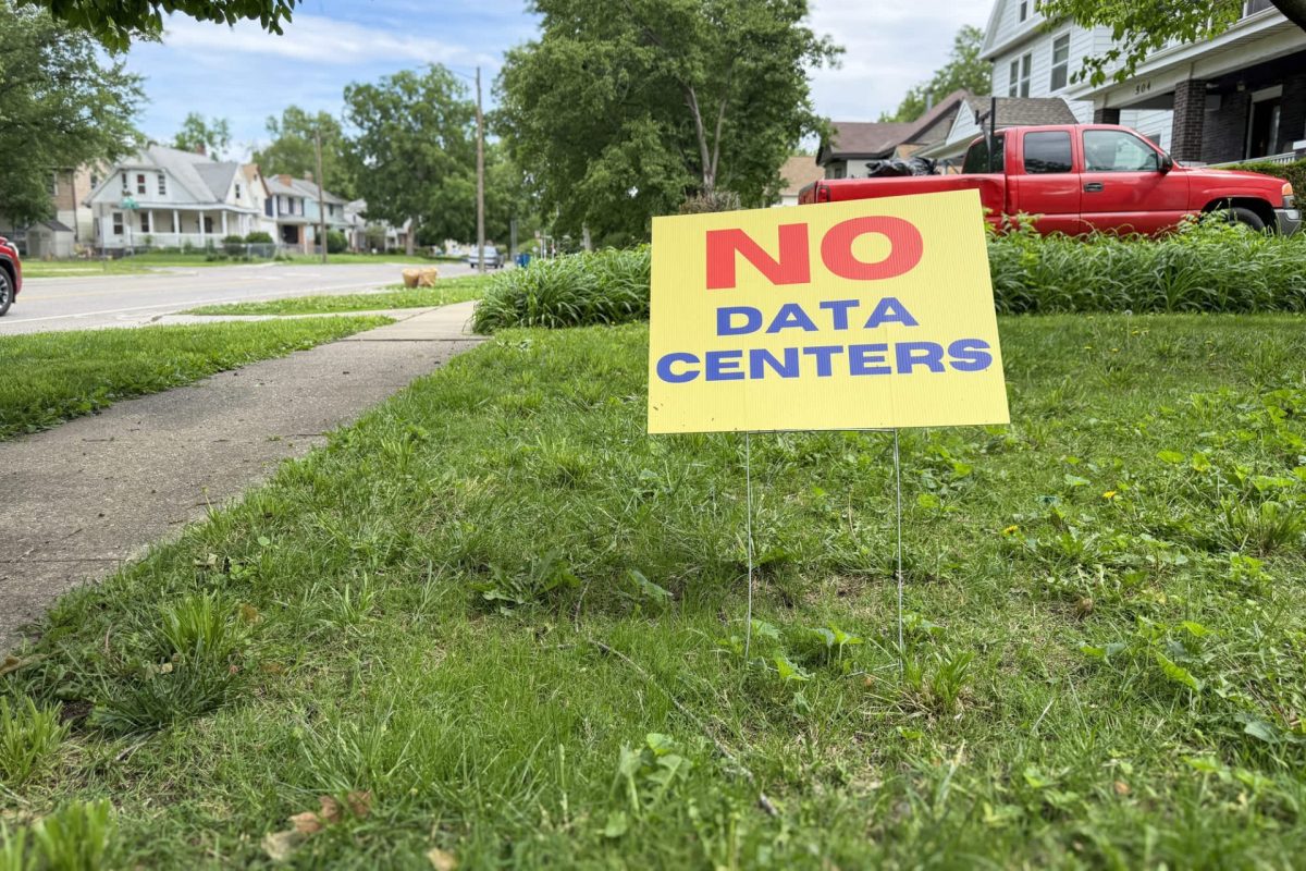 A bright yellow sign reading "NO DATA CENTERS" stands in a grassy yard next to a sidewalk, with suburban homes in the background.