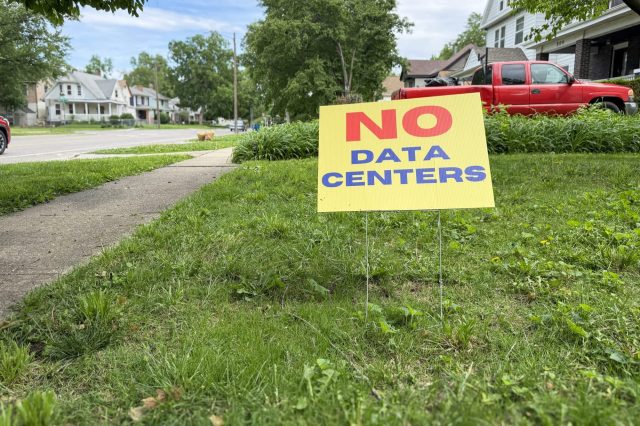 A bright yellow sign reading "NO DATA CENTERS" stands in a grassy yard next to a sidewalk, with suburban homes in the background.