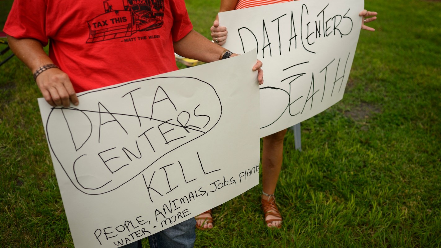 Two individuals hold protest signs criticizing data centers, stating they "kill" and cause harm to people and nature.