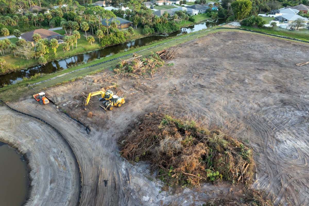 Construction machinery clearing land near a residential area with a waterway in the background.