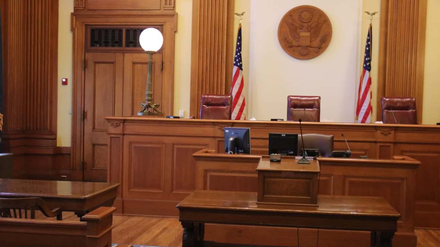 A wooden courtroom with two flags, a seal, and empty chairs behind a judge's bench.