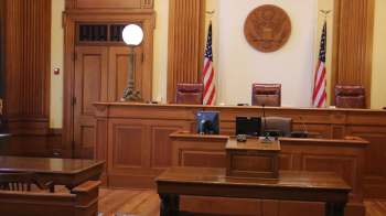 A wooden courtroom with two flags, a seal, and empty chairs behind a judge's bench.
