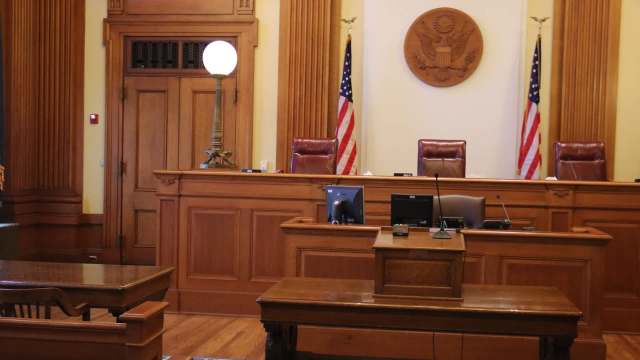 A wooden courtroom with two flags, a seal, and empty chairs behind a judge's bench.
