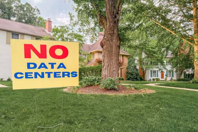 A bright yellow sign in a yard reads "NO DATA CENTERS," surrounded by lush greenery and residential homes.