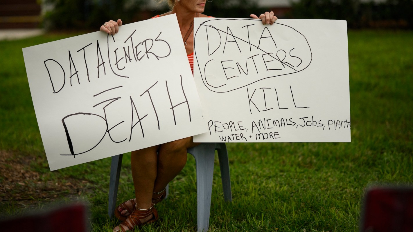 A person holding protest signs that read "Data Centers Kill" and "Data Centers Death," expressing concerns for the environment.