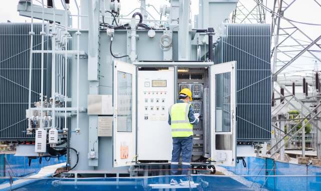 A technician in a helmet and vest inspects equipment at an electrical substation with control panels open.