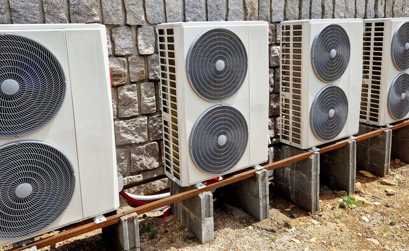 Multiple outdoor air conditioning units are mounted on concrete blocks against a stone wall.