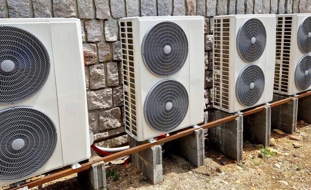Multiple outdoor air conditioning units are mounted on concrete blocks against a stone wall.