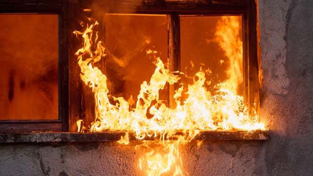 Flames engulf the window of a building, illuminating the interior with a fiery glow.