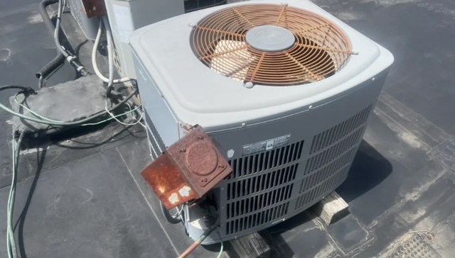 An outdoor air conditioning unit sits on a flat rooftop, with visible electrical connections and rusted components.