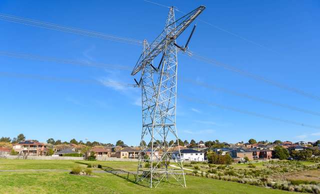 A tall electricity transmission tower stands against a clear blue sky near a suburban neighborhood.
