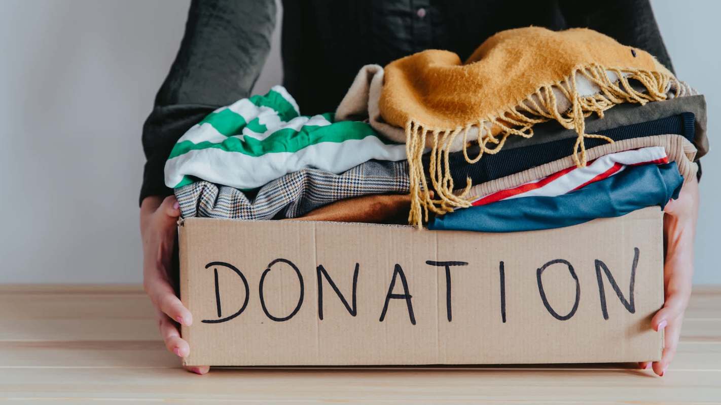 A person holds a cardboard box labeled "DONATION" filled with various clothing items.