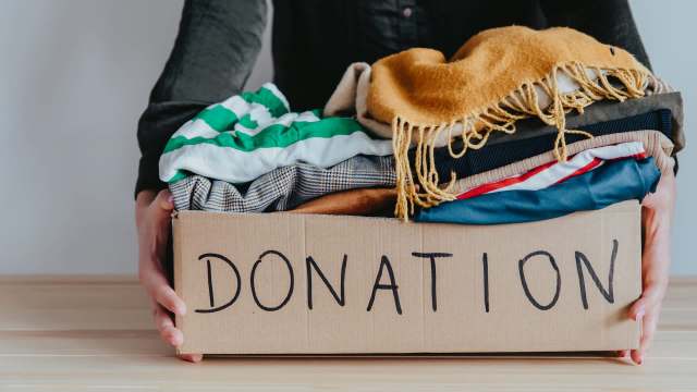 A person holds a cardboard box labeled "DONATION" filled with various clothing items.