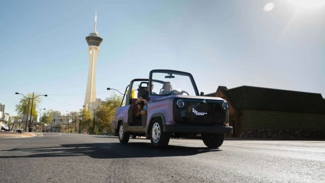 A convertible car drives down a city street with a tall tower in the background under a clear blue sky.