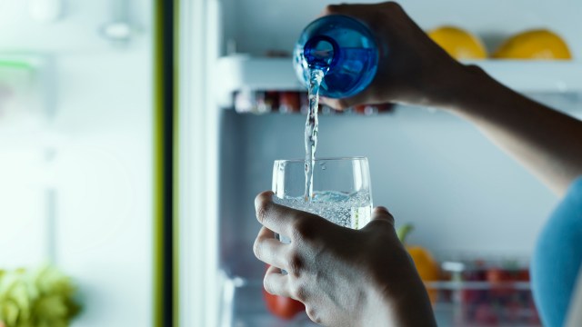 A person pours sparkling water from a blue bottle into a clear glass inside a refrigerator.