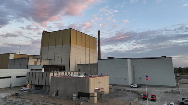 A view of industrial buildings with a smokestack under a cloudy sky, featuring parked vehicles and an American flag.
