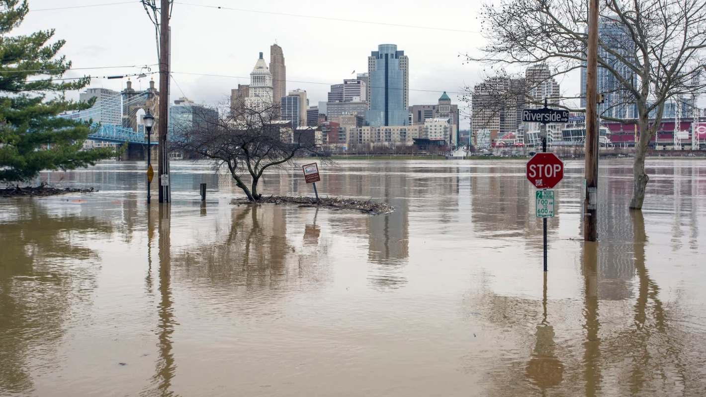 The Cincinnati city skyline partially submerged in floodwaters with trees and street signs visible above the water.