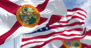 The Florida state flag waving with the American flag in the background against a blue sky.